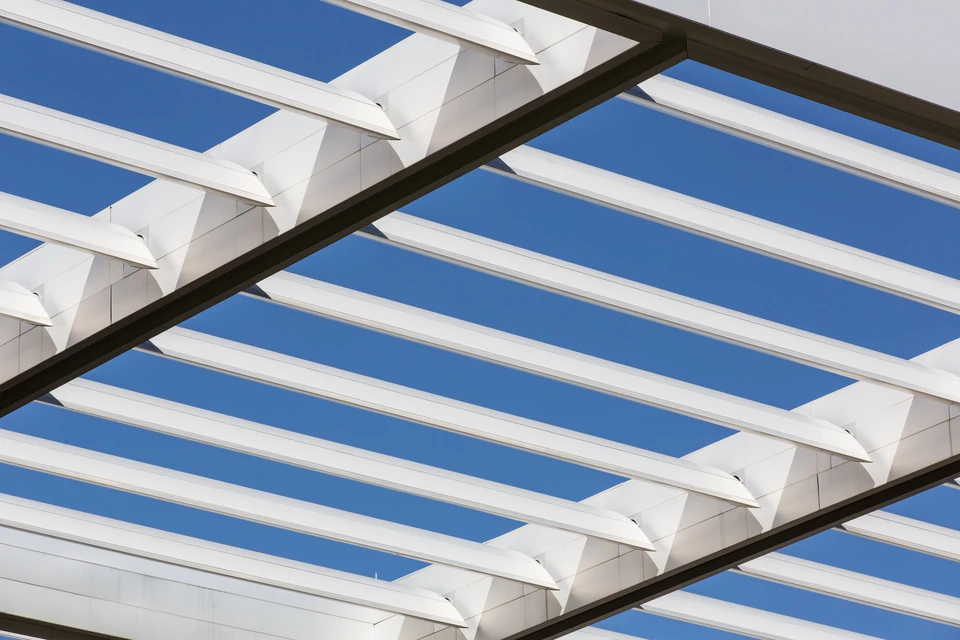 Scène extérieure montrant une pergola moderne en cours de construction ou terminée, photographiée en contre-plongée sous un ciel bleu dégagé
